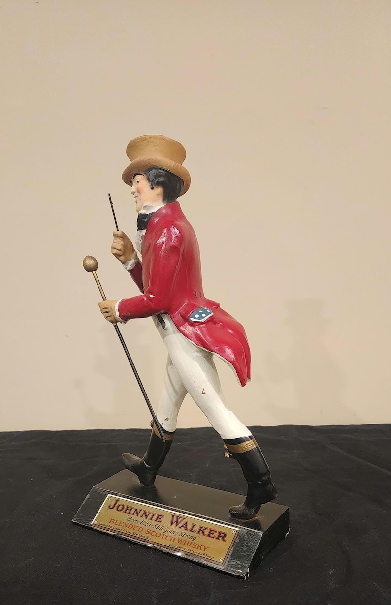 K&ouml;nnte beinhalten: Figur des Johnnie Walker Striding Man. Die Figur tr&auml;gt einen roten Mantel, eine wei&szlig;e Hose und schwarze Stiefel und h&auml;lt einen Stock und einen kleinen Stab. Auf dem Sockel stehen der Markenname und Text.
