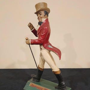 Puede incluir: Figura decorativa de un hombre con abrigo rojo, pantalones blancos y botas negras, sosteniendo un bast&oacute;n. La figura est&aacute; sobre una base verde con el texto "JOHNNIE WALKER". El hombre lleva un sombrero de copa.