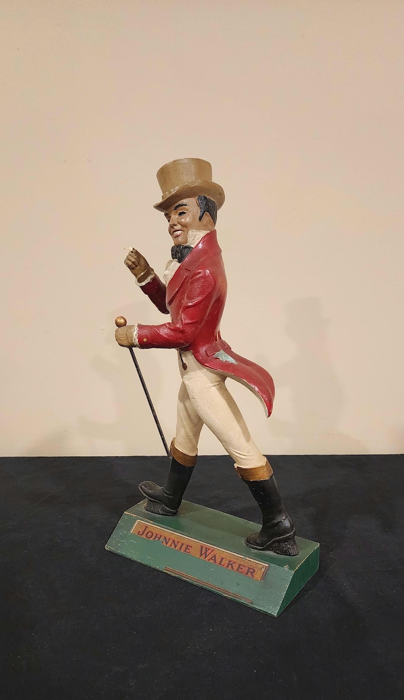 Puede incluir: Figura decorativa de un hombre con abrigo rojo, pantalones blancos y botas negras, sosteniendo un bast&oacute;n. La figura est&aacute; sobre una base verde con el texto "JOHNNIE WALKER". El hombre lleva un sombrero de copa.