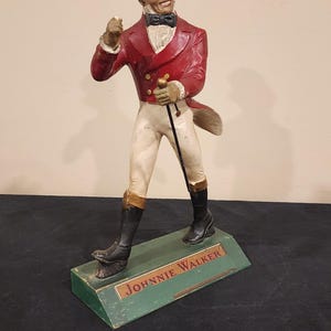 Puede incluir: Una figura vintage de Johnnie Walker. La figura viste una chaqueta roja, pantalones blancos y un sombrero de copa, sosteniendo un bast&oacute;n. La base es verde con el nombre de la marca.