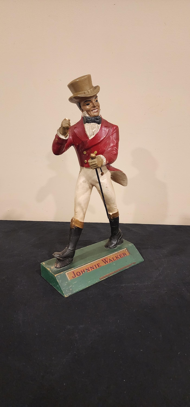 Puede incluir: Una figura vintage de Johnnie Walker. La figura viste una chaqueta roja, pantalones blancos y un sombrero de copa, sosteniendo un bast&oacute;n. La base es verde con el nombre de la marca.