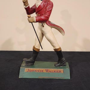Puede incluir: Una figura vintage de Johnnie Walker. La figura viste un abrigo rojo, pantalones blancos y botas negras, sosteniendo un bast&oacute;n. La base es verde con el nombre de la marca.
