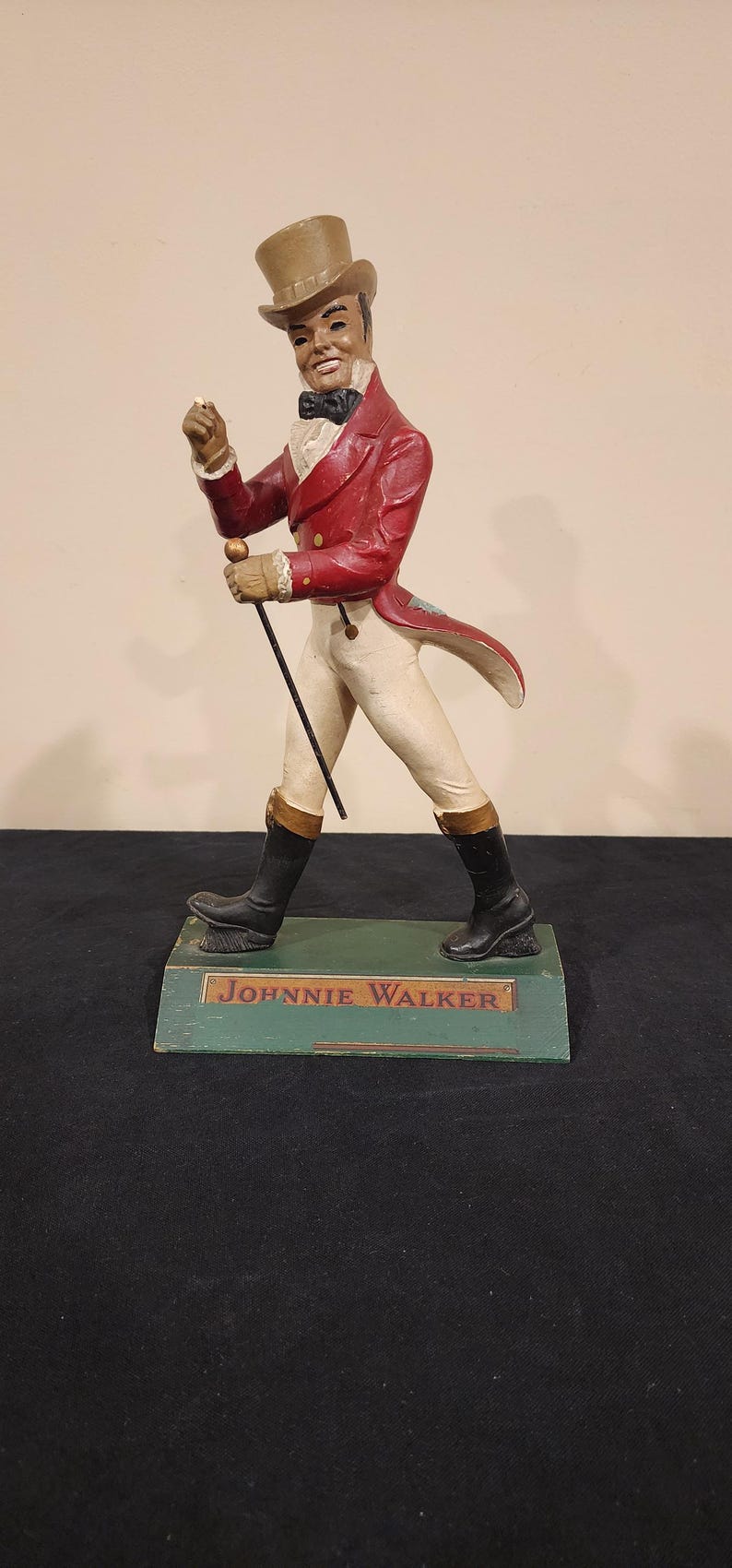 Puede incluir: Una figura vintage de Johnnie Walker. La figura viste un abrigo rojo, pantalones blancos y botas negras, sosteniendo un bast&oacute;n. La base es verde con el nombre de la marca.