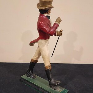 Puede incluir: Figura vintage de un caballero con levita roja, pantalones blancos y botas negras, sosteniendo un bast&oacute;n. La figura lleva un sombrero de copa y guantes, de pie sobre una base verde. Una pieza decorativa cl&aacute;sica.