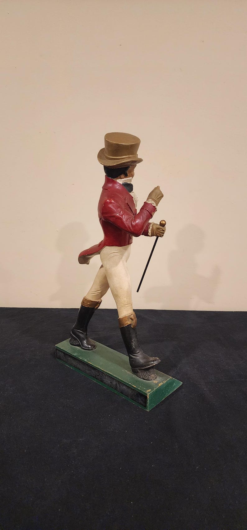 Puede incluir: Figura vintage de un caballero con levita roja, pantalones blancos y botas negras, sosteniendo un bast&oacute;n. La figura lleva un sombrero de copa y guantes, de pie sobre una base verde. Una pieza decorativa cl&aacute;sica.