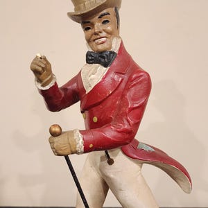 Puede incluir: Figura vintage de un hombre con abrigo rojo, pantalones blancos y botas negras, sosteniendo un bast&oacute;n y un objeto peque&ntilde;o. Lleva un sombrero de copa y una pajarita. La base dice "JOHNNIE WALKER".