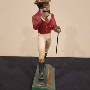 Puede incluir: Figura vintage de un hombre con chaqueta roja, pantalones blancos y botas negras, sosteniendo un bast&oacute;n. La figura lleva un sombrero de copa y est&aacute; sobre una base verde con el texto "Johnnie Walker".