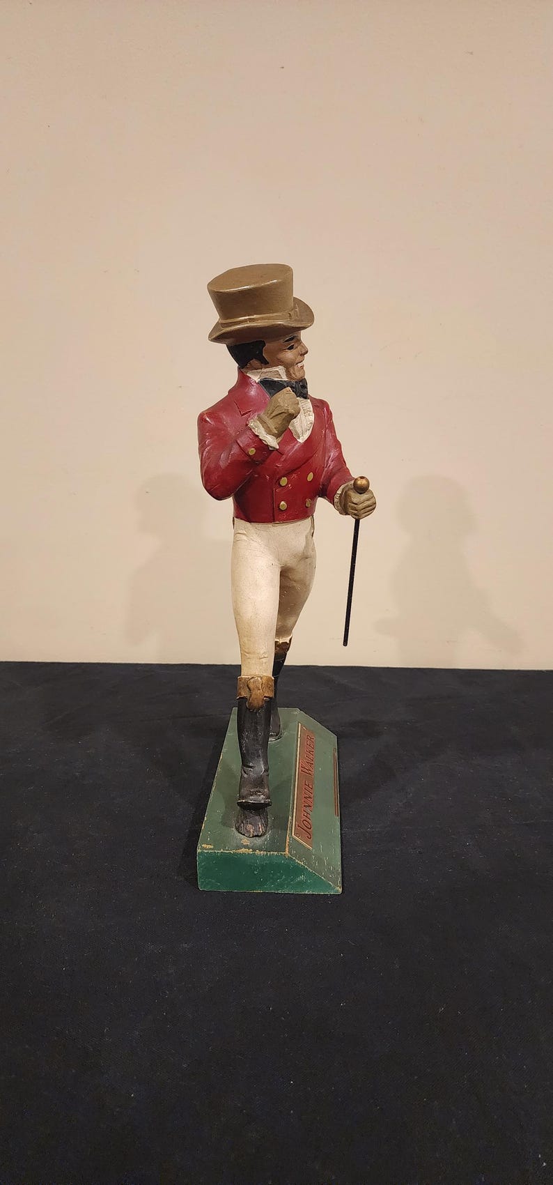 Puede incluir: Figura vintage de un hombre con chaqueta roja, pantalones blancos y botas negras, sosteniendo un bast&oacute;n. La figura lleva un sombrero de copa y est&aacute; sobre una base verde con el texto "Johnnie Walker".