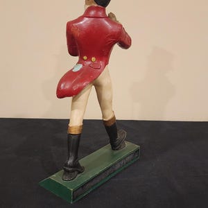 Puede incluir: Figura decorativa de un hombre con abrigo rojo, pantalones beiges y botas negras, con un sombrero de copa marr&oacute;n. La figura est&aacute; sobre una base verde. La figura est&aacute; de espaldas al espectador.