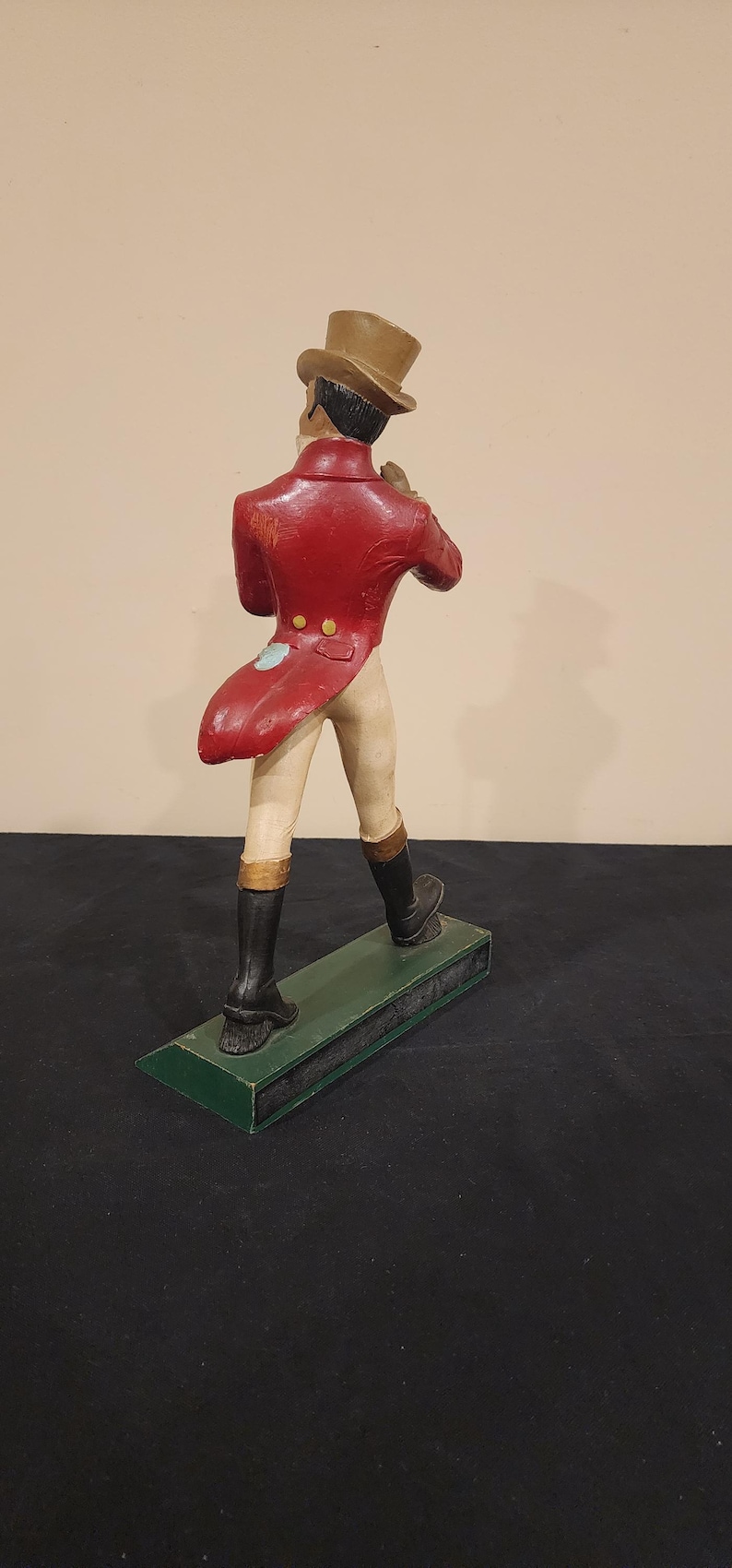 Puede incluir: Figura decorativa de un hombre con abrigo rojo, pantalones beiges y botas negras, con un sombrero de copa marr&oacute;n. La figura est&aacute; sobre una base verde. La figura est&aacute; de espaldas al espectador.