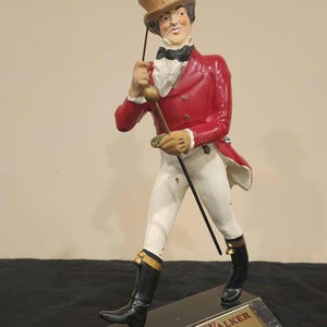 K&ouml;nnte beinhalten: Eine Figur von Johnnie Walker, einem Mann in einem roten Mantel, wei&szlig;en Hosen und schwarzen Stiefeln, der einen Stock h&auml;lt und einen Zylinder tr&auml;gt. Der Sockel hat eine Plakette mit der Aufschrift "Johnnie Walker Blended Scotch Whisky."