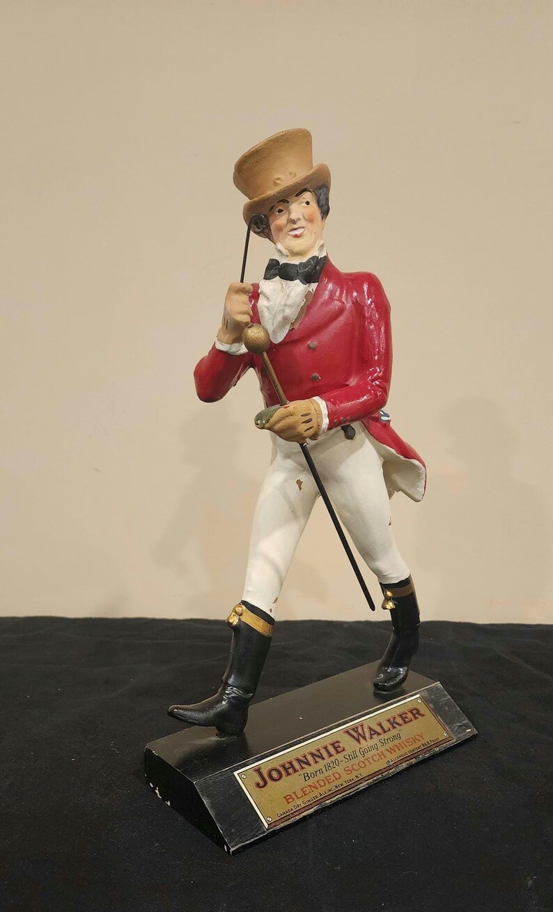 K&ouml;nnte beinhalten: Eine Figur von Johnnie Walker, einem Mann in einem roten Mantel, wei&szlig;en Hosen und schwarzen Stiefeln, der einen Stock h&auml;lt und einen Zylinder tr&auml;gt. Der Sockel hat eine Plakette mit der Aufschrift "Johnnie Walker Blended Scotch Whisky."