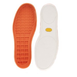 Op de afbeelding: Close-up van de onderkant van een paar sneakers. De linker schoen heeft een oranje rubberen zool met een rasterpatroon. De rechter schoen heeft een witte rubberen zool met een textuurpatroon en een geel Vibram-logo.