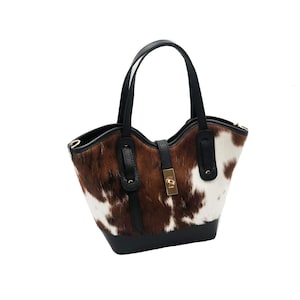 Op de afbeelding: Een tote bag van koeienhuid met een zwarte basis, handvatten en afwerking. De tas heeft een bruin-wit gevlekt patroon, een goudkleurige sluiting en een gestructureerd ontwerp. De handvatten zijn gebogen en gemaakt van zwart leer.
