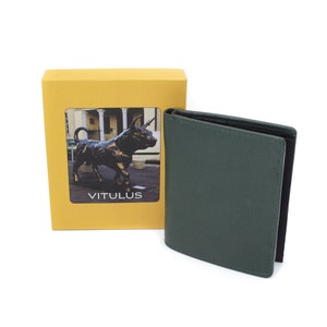May include: A green leather wallet with a black interior. The wallet is closed and sitting on a white surface. The wallet is next to a yellow box with a picture of a bronze bull statue and the word "VITULUS" printed on it.