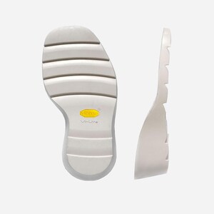 May include: A white shoe sole featuring horizontal tread grooves and a yellow Vibram Vi-Lite logo. The sole is shown from both a bottom view and a side profile view, highlighting the thick, lightweight material and the textured grip pattern.