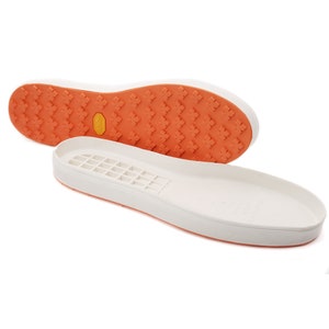 Op de afbeelding: Een paar witte schoenzool met een getextureerde oranje buitenzool. De buitenzool heeft een patroon van kleine kruisen en de tekst "Vibram" is zichtbaar.