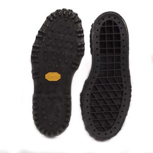 Peut inclure: Deux semelles de bottes en caoutchouc noir avec des motifs de bande de roulement texturés. L'une présente un design à crampons, l'autre un motif quadrillé. Un logo "Vibram" jaune est visible sur une semelle.