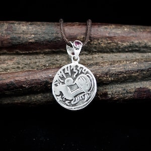 May include: A silver pendant necklace with a circular charm featuring a detailed design. The charm is suspended from a brown cord and has a small, round, purple gemstone at the top. The pendant rests on a piece of dark brown wood against a black background.