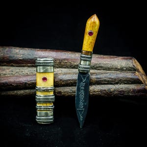 May include: A small knife with a dark metal blade featuring etched symbols, a light-coloured textured handle with a red stone inlay, and a matching cylindrical sheath with silver-toned metal bands and a central red stone inlay.