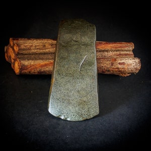 May include: A green stone hand axe with a rough, textured surface. The axe is resting on top of a stack of brown wooden logs.