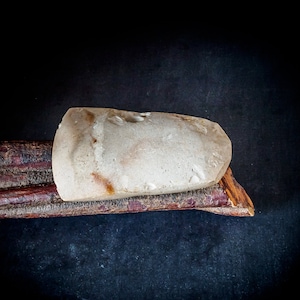 以下が含まれることがあります： 赤みがかった筋が入った濃い茶色の木の上に置かれた、薄い茶色で滑らかで楕円形の石