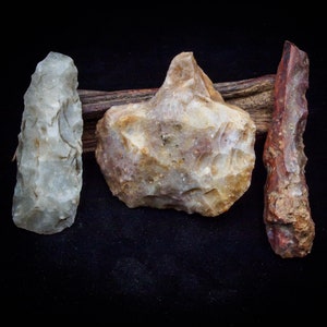 May include: Three flint arrowheads, one with a light gray color, one with a brown and white color, and one with a red and brown color, are displayed on a dark background.
