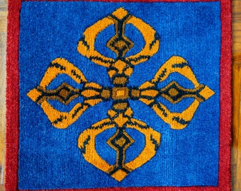 Peut inclure: Un tapis bleu avec une bordure rouge présente un grand symbole orange, noir et marron au centre. Le symbole est un motif stylisé d'une croix à quatre boucles.