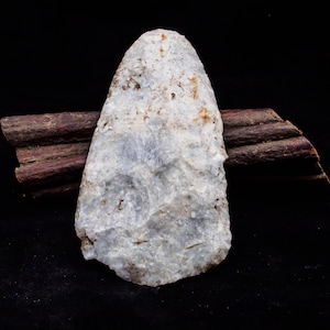 May include: A white, opaque, arrowhead-shaped stone tool with a rough, textured surface. The tool is resting on a piece of brown wood.