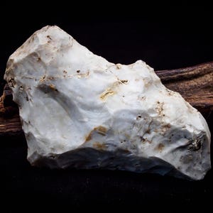 May include: A white, irregularly shaped stone with a rough, textured surface. The stone has a light brown color in some areas. It is resting on a piece of dark brown wood.