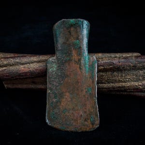 May include: A bronze axe head with a wooden handle. The axe head has a rectangular blade and a handle that appears to be made of a piece of wood. The bronze has a green patina, and the wood is brown.
