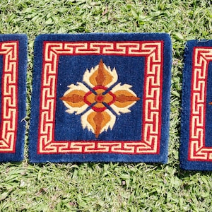 May include: Three square, navy blue decorative cushions with a central floral design in shades of orange, tan, and cream. Each cushion has a red and tan border with a repeating geometric pattern. The cushions are displayed on a bed of green grass.