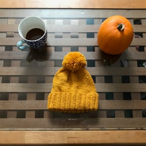 May include: A yellow knitted beanie with a fluffy pom pom on top. The beanie is sitting on a wooden table with a cup of coffee and a pumpkin.
