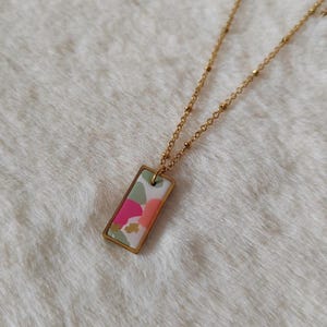 May include: A gold-tone necklace with a rectangular pendant. The pendant displays a colourful abstract design with pink, green, and orange shapes on a white background. The necklace chain has small, round beads.