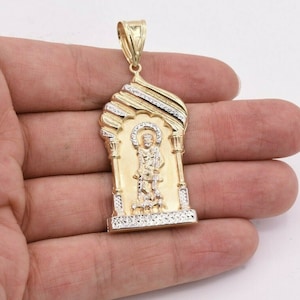 May include: Gold-tone pendant featuring a religious figure within an arched frame. The pendant has a detailed design with a halo and intricate detailing. The frame has a textured design with white accents. The bail is also gold-toned.