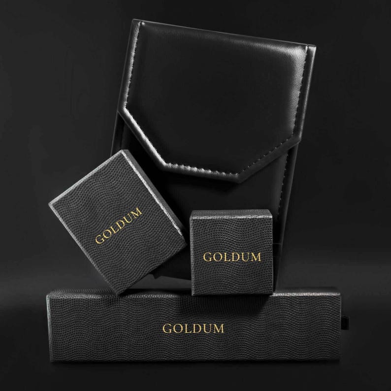 Puede incluir: Un conjunto de cajas de joyer&iacute;a negras y un estuche de cuero. Las cajas tienen una textura y presentan el texto dorado "GOLDUM". La disposici&oacute;n sugiere una presentaci&oacute;n o almacenamiento, con enfoque en el lujo y la elegancia.