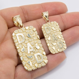 Op de afbeelding: Twee gouden hangers met het woord "DAD" in reliëfletters. De hangers zijn rechthoekig en hebben een getextureerd oppervlak.