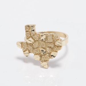 May include: A gold ring in the shape of Texas. The ring has a textured, nugget-style design on the Texas outline and a plain band. The ring is set against a white background.