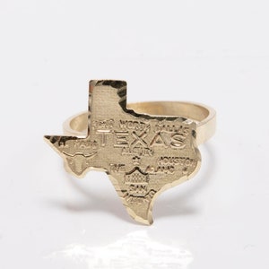 May include: A gold-colored ring shaped like the state of Texas. The ring features the word "TEXAS" and the names of several cities within the state. The ring is on a white background.