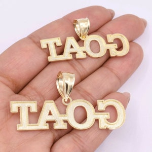 May include: Two gold pendants with the word "GOAT" spelled out in block letters. The pendants are on a hand with the word "GOAT" facing the camera.