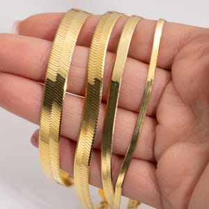 May include: A hand displays several gold-colored herringbone chain necklaces of varying widths. The chains have a flat, flexible design with a subtle, textured pattern. The necklaces are displayed against a neutral background.