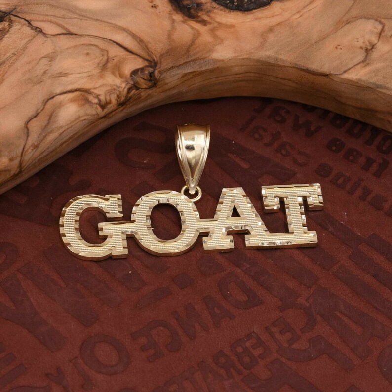 May include: Gold pendant necklace with the word "GOAT" spelled out in block letters. The letters have a diamond-cut texture.