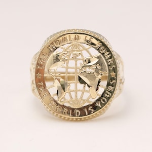 May include: A gold ring featuring a circular face with a cut-out globe design. The outer border of the circle is engraved with the text THE WORLD IS YOURS, separated by two small stars. The ring has a polished finish with textured details.