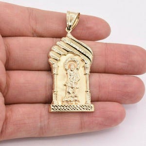 May include: A gold-colored pendant featuring a religious figure within an arched frame. The pendant has intricate detailing, including columns and a patterned base. The bail is a simple loop for a chain. The overall design is ornate and detailed.