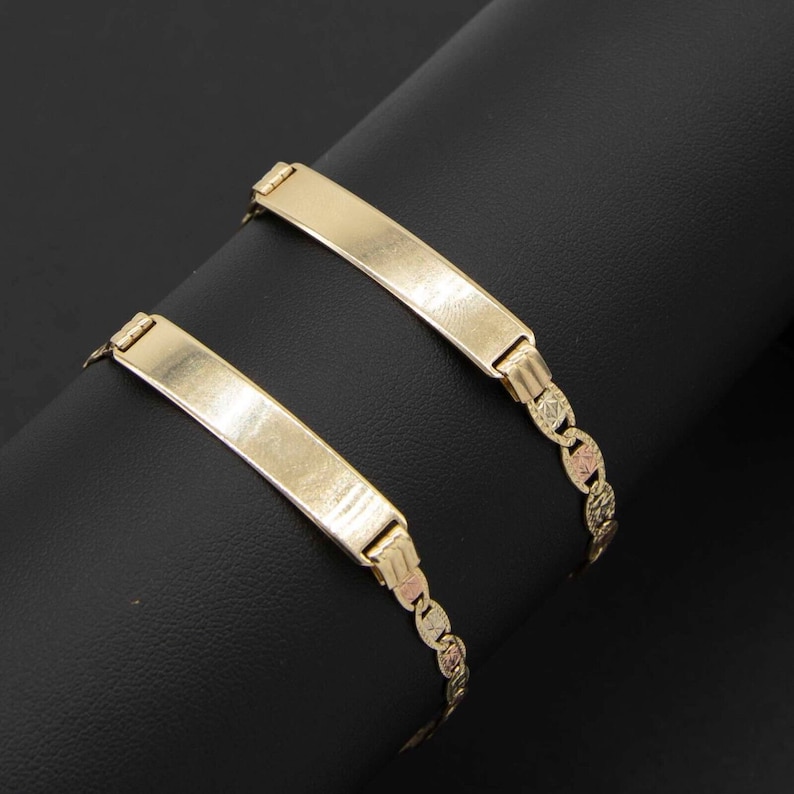 May include: Two gold bracelets with a flat, rectangular plate on each. The plates are blank and can be engraved. The bracelets are on a black surface.
