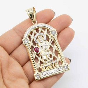 May include: Gold and silver pendant featuring a depiction of San Lazaro, a Catholic saint, with a red gemstone. The pendant is surrounded by a decorative latticework design and the words "SAN LAZARO" are inscribed at the bottom.