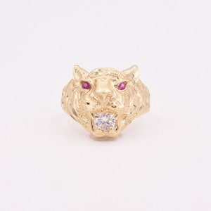 Op de afbeelding: Een gouden ring met een tijgerkopontwerp. De tijger heeft rode ogen en een enkele witte steen in zijn bek.