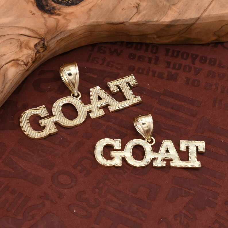 May include: Two gold-tone pendants spelling out the word "GOAT" in a diamond-cut style. The pendants are on a brown textured surface.