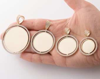 May include: Four gold-tone pendants with a round, clear, blank center surrounded by a circle of clear stones. The pendants are of different sizes and are displayed on a hand.