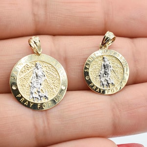 May include: Two gold-tone Saint Lazarus pendants. Each circular pendant features a detailed depiction of Saint Lazarus in silver-tone metal, surrounded by the words "SAINT LAZARUS" and "PRAY FOR US" in gold lettering. The pendants have a textured background.